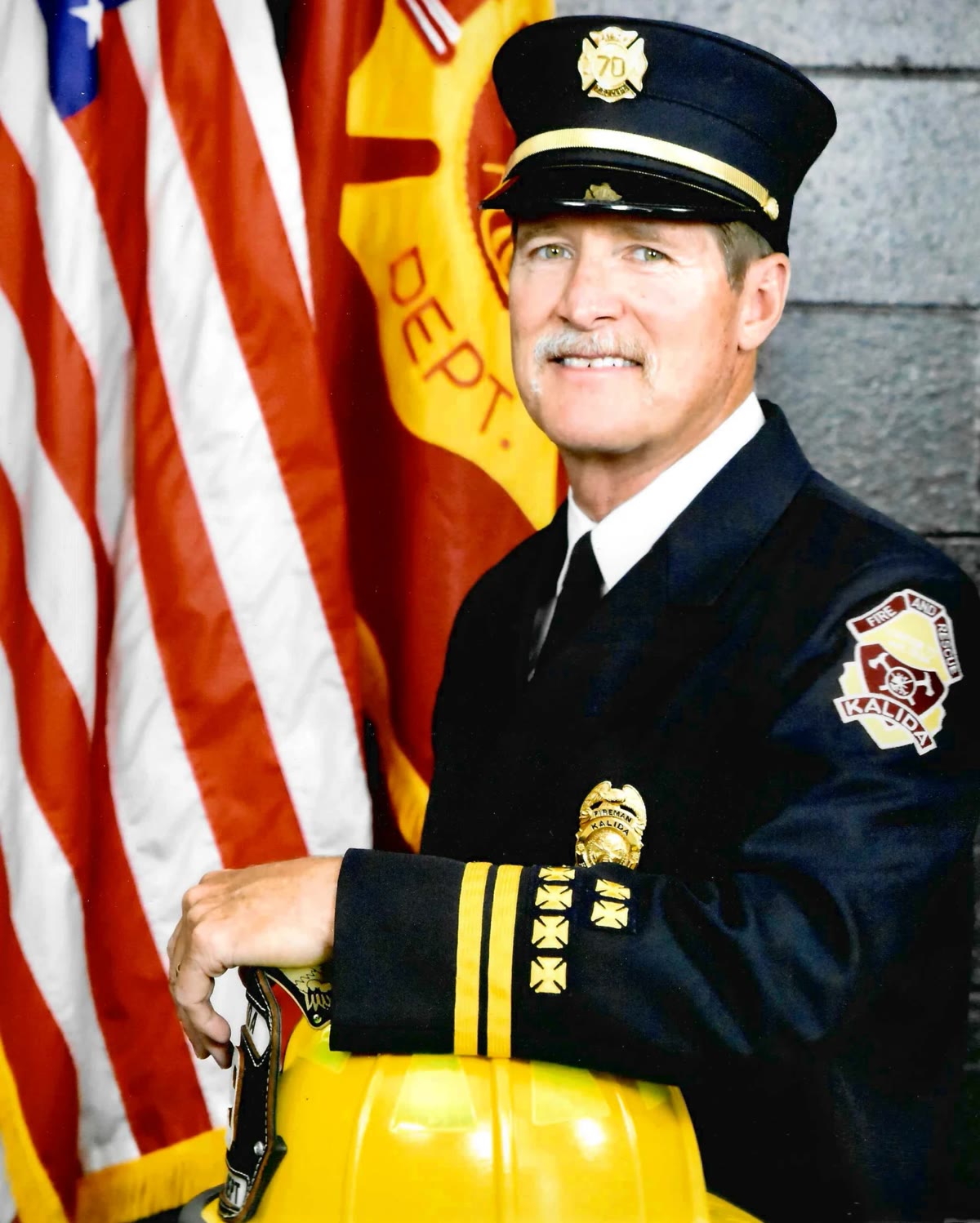 Brian Berheide smiling in his Kalida Fire Department dress uniform, holding his helmet in front of the American flag
